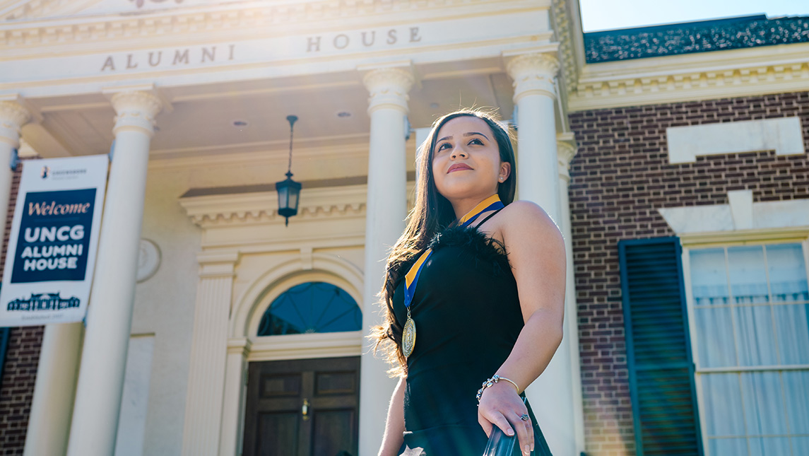 Young woman in a dress with a medal around her neck poses in front of Alumni House.