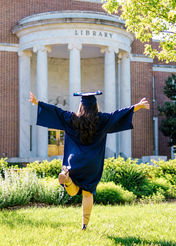 We see a woman in cap and gown from behind her arms are outstretched & her heel is kicked up in celebration. She's running towards Jackson Library.