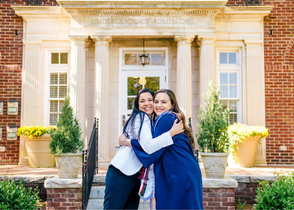 A woman hugs a young woman in a graduation gown in front of the Admissions Building.