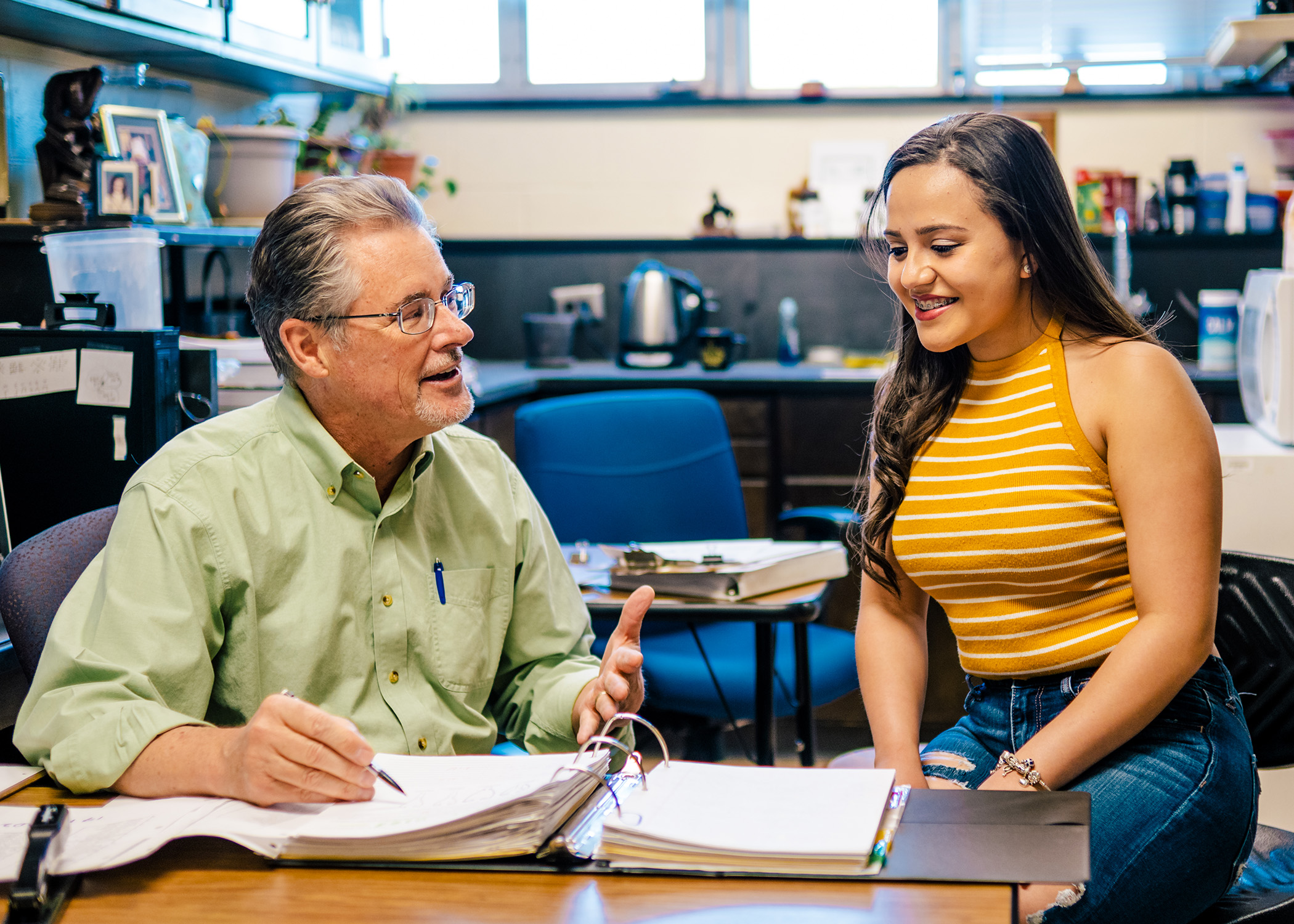 Smiling young woman sits in an academic office with a professor who gestures to a notebook.