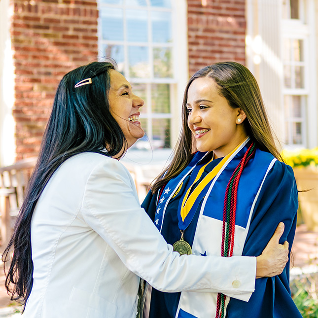 Woman smiles with pride as she hugs a young woman in commencement gown with honors cords. 