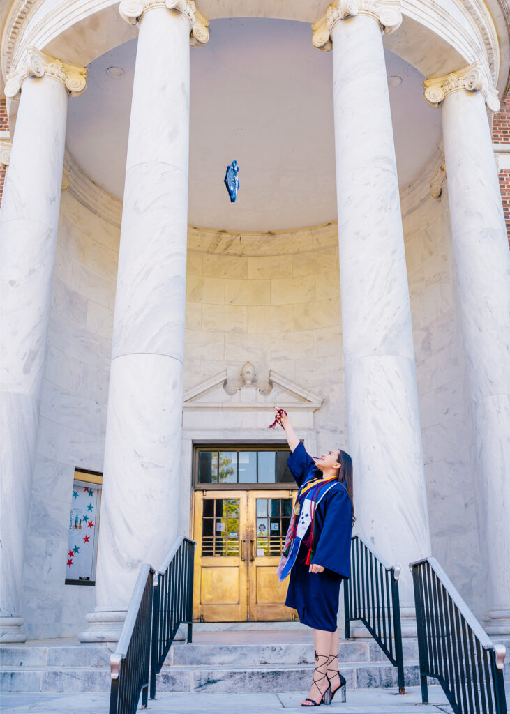 Woman in graduation gown stands on steps of Jackson Library and throws her cap in the air.