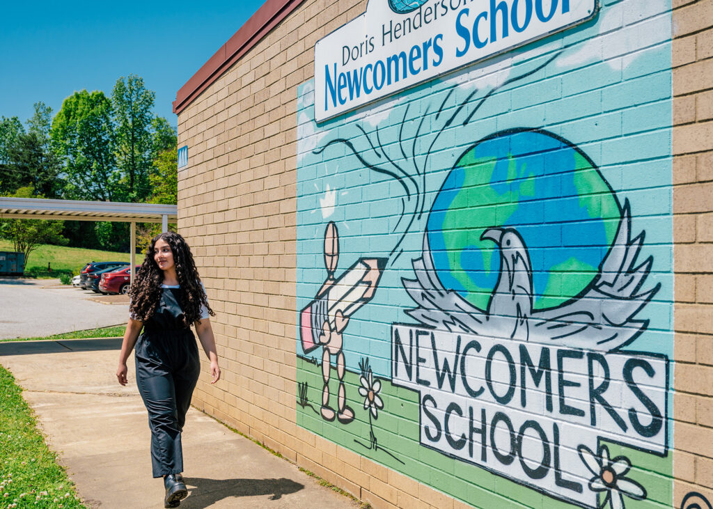 Young woman walks down a sidewalk in front of a brick wall with Newcomers School sign and graffiti of a pencil and globe.