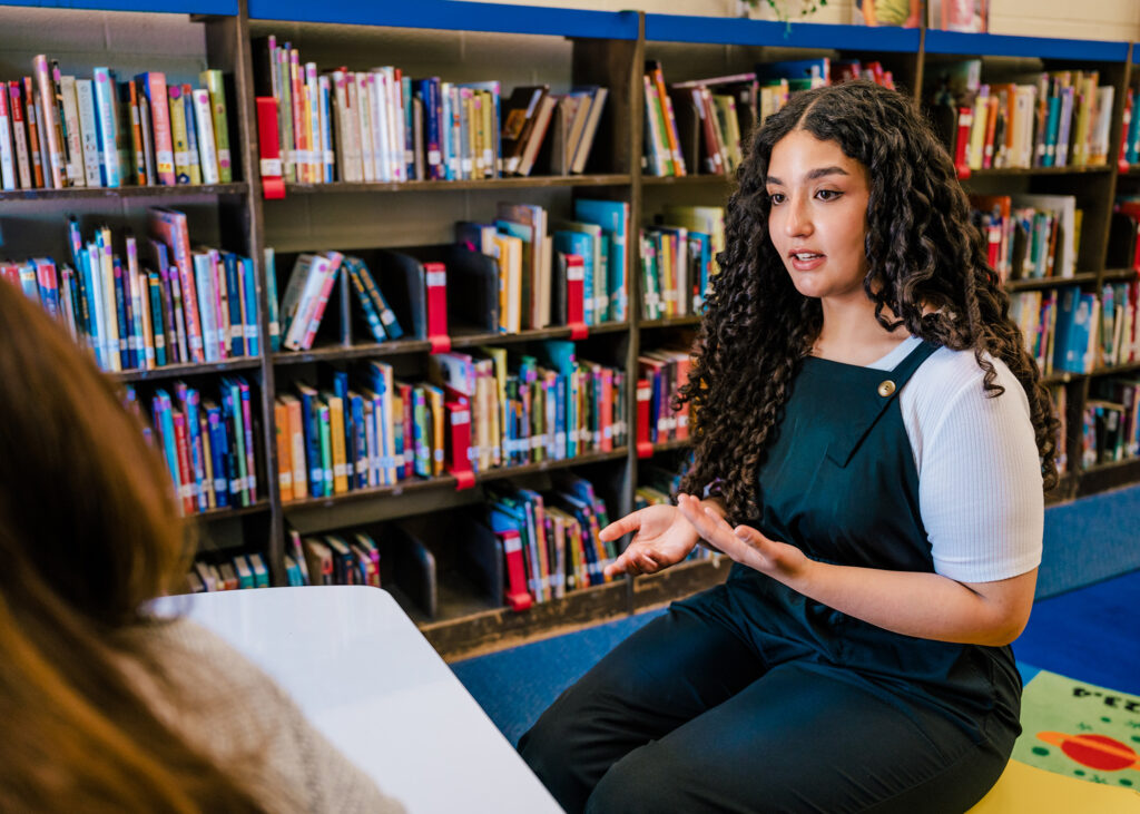 Young woman sits in a library talking to someone in the foreground. 