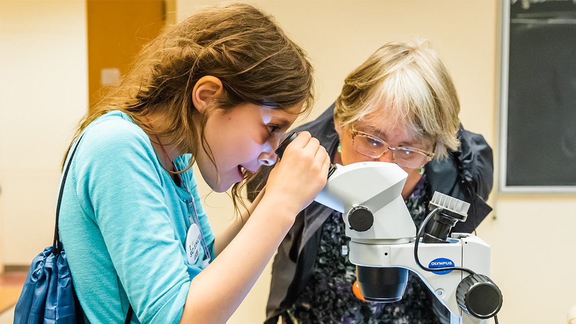 Young girl looks in a microscope while an older lady stands behind.