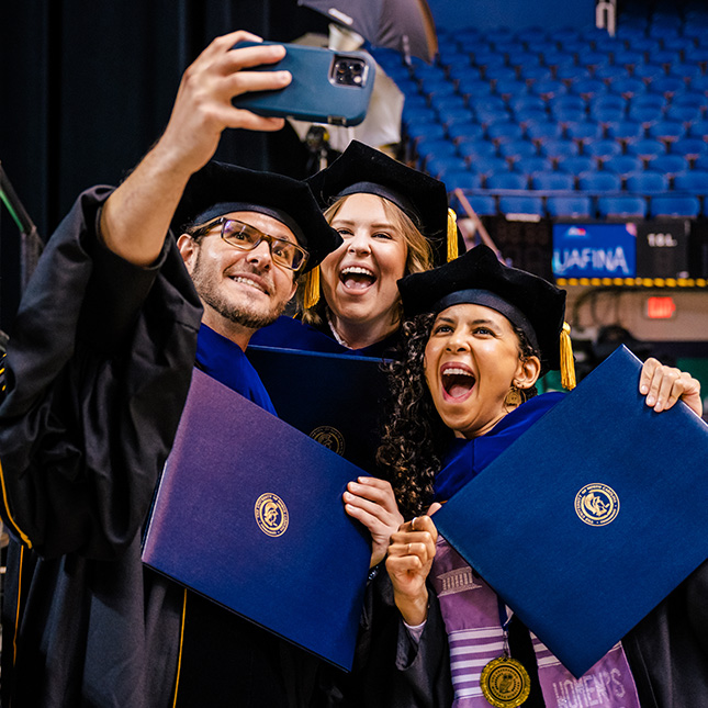Three masters graduates pose for a selfie in cap and gown.