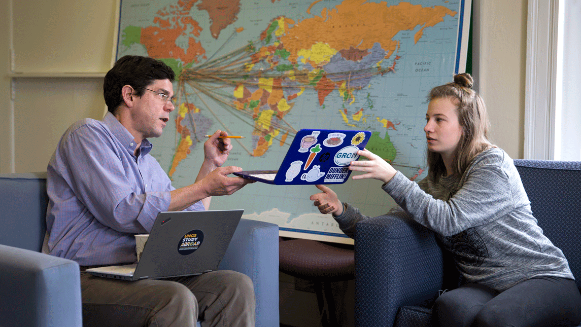 A student hands a professor her laptop in front of a world map