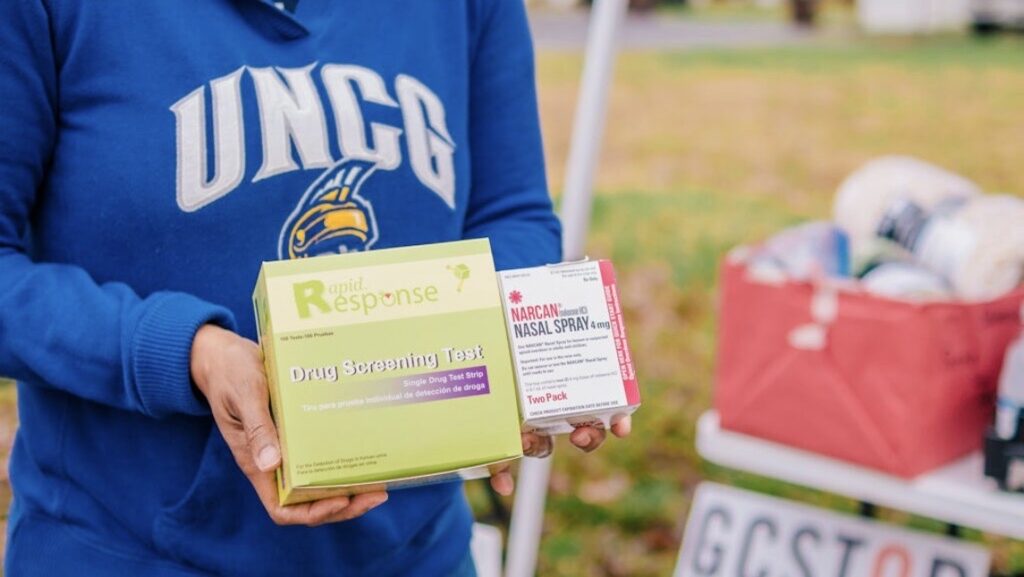 Someone in a UNCG sweatshirt holds 2 boxes - drug screening & nasal Naloxone at a GCSTOP distribution tent.
