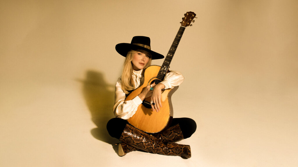 Singer Jewel sits on the ground with her legs crossed. A guitar sits in her lap.