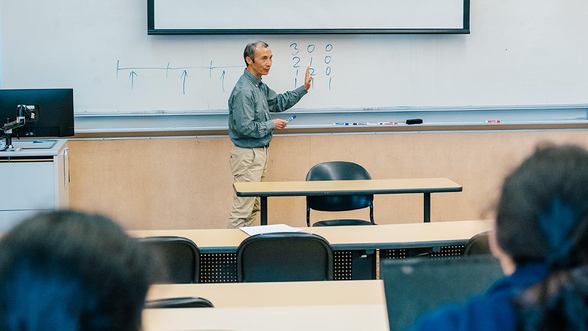 Professor writes on white board at the head of a class.