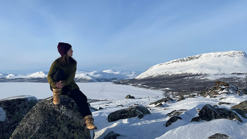 Woman in hiking boots sits on a rock and looks to the horizon with a snowy mountain terrain in the background.