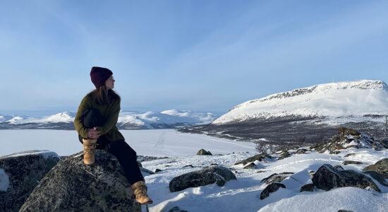 Woman in hiking boots sits on a rock and looks to the horizon with a snowy mountain terrain in the background.
