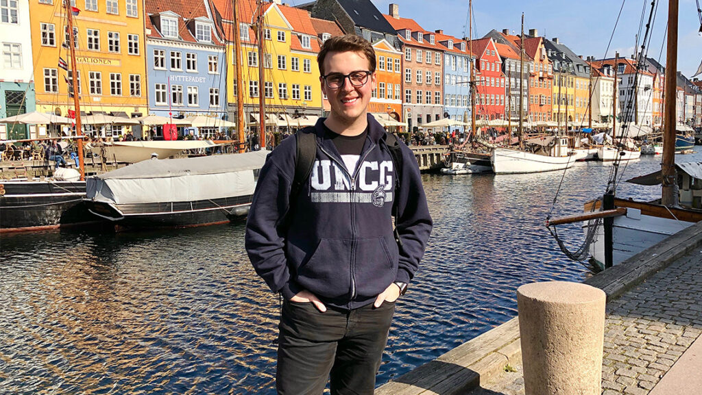 Student in UNCG jacket smiles at camera in front of colorful buildings in Germany