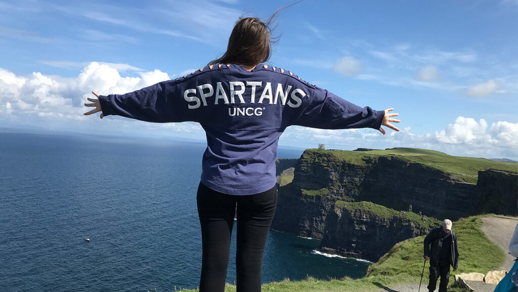 Woman with shirt that states "Spartans -UNCG " opening her arms overlooking a majestic view of land and water