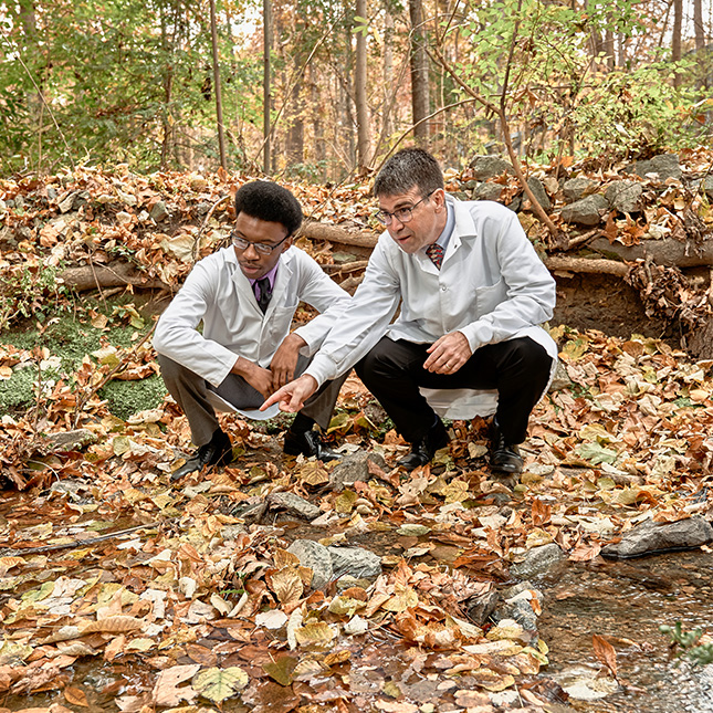 A student and professor in white lab coats crouch near a creekbed in the woods collecting water samples.