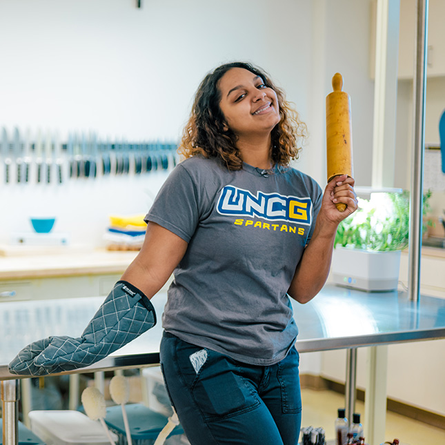Female student in UNCG t-shirt holds a rolling pin and oven mit in a kitchen lab.