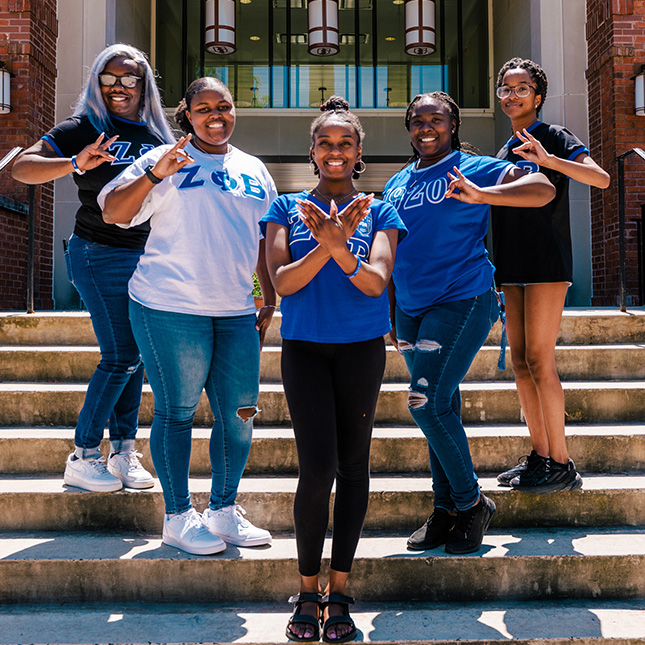 5 girls wearing t-shirts with greek letters stand on the steps of a UNCG campus building.