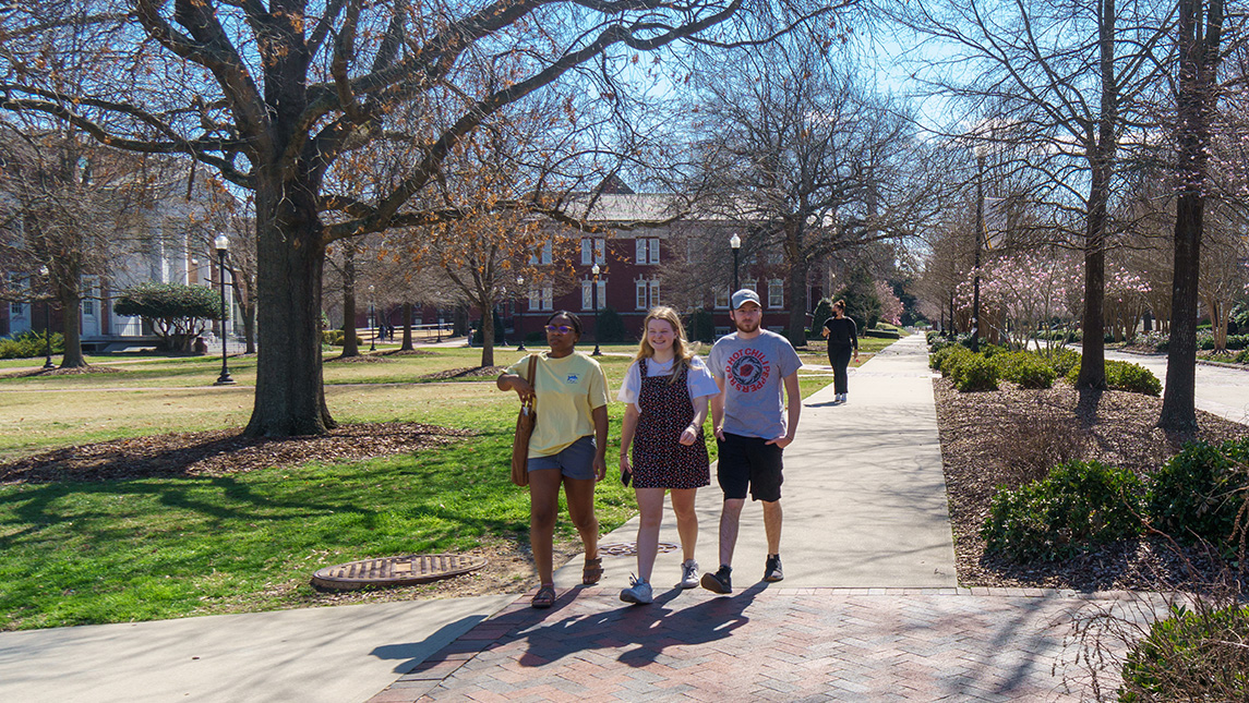 Three students walking on campus sidewalks with trees and buildings in the background.