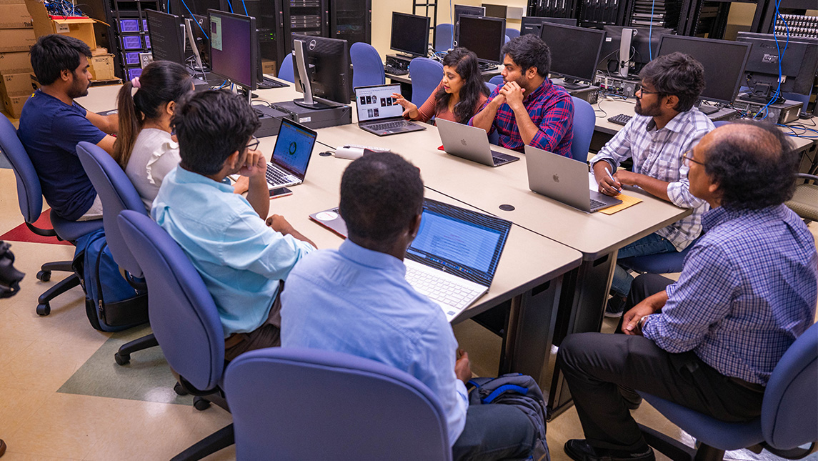 computer science students seated around tables together, all with laptops open and having a discussion.