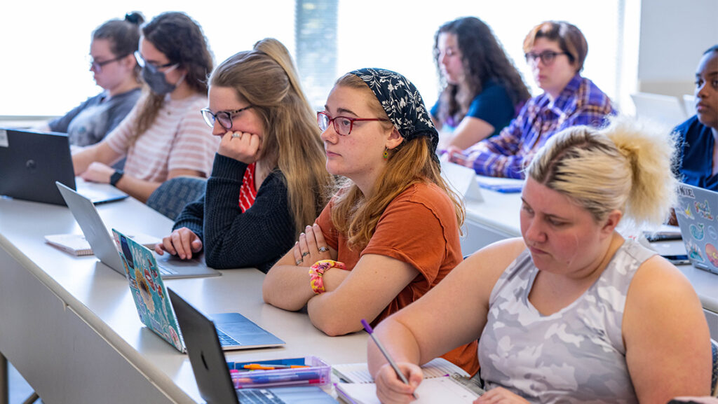 UNCG students sit at a classroom desk taking notes on their laptops or notebooks.