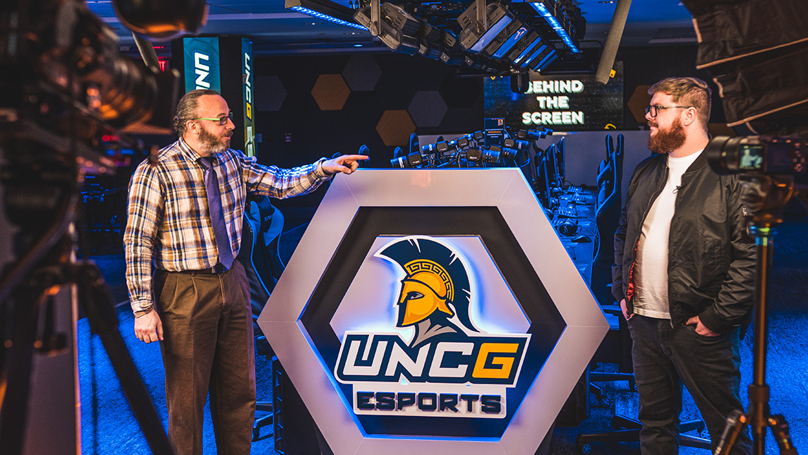 2 men talk in the esports arena with the UNCG logo between them and cameras set up around.