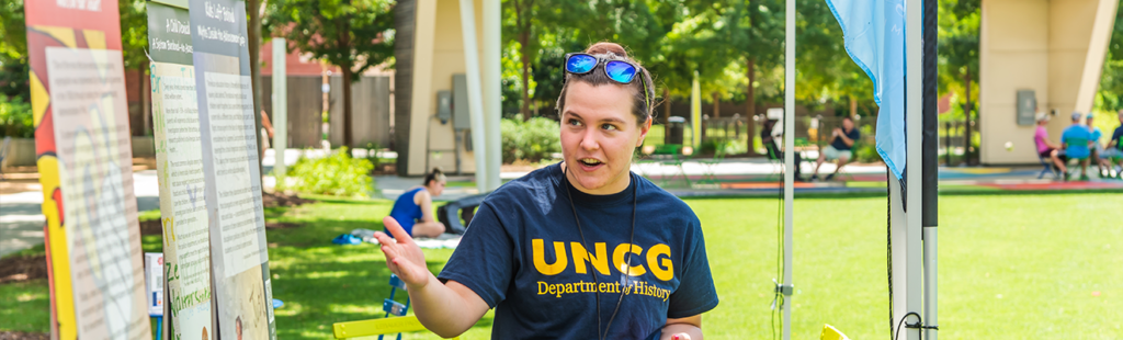 female student wearing UNCG History shirt points at informational banners in a park