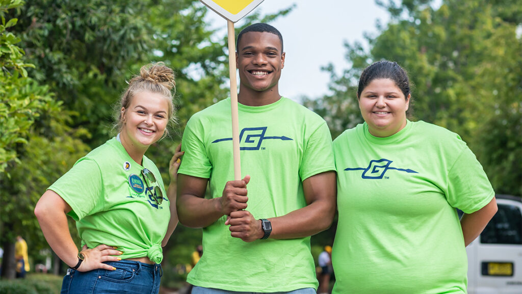 Three people in green UNCG SPEAR t-shirts hold up a sign welcoming new students.