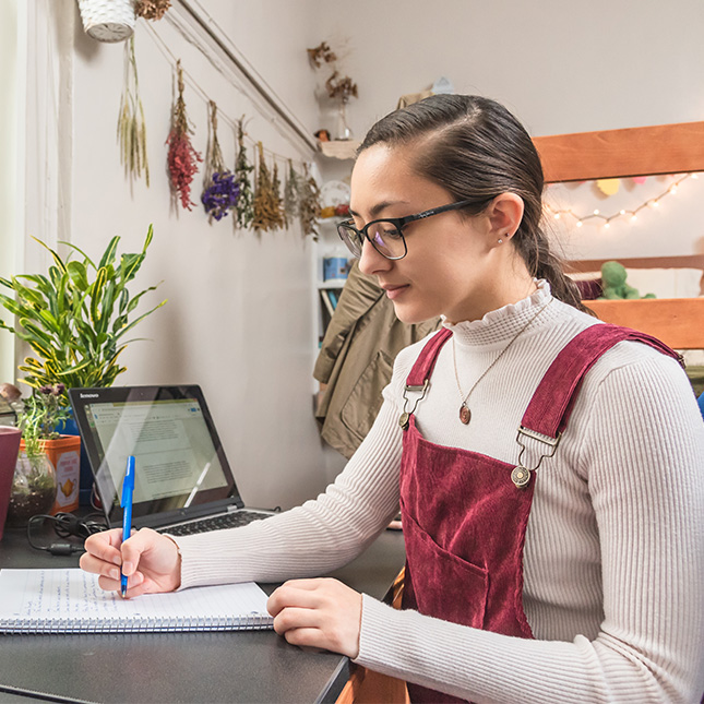 Student in room of residence hall takes notes at desk with laptop. 