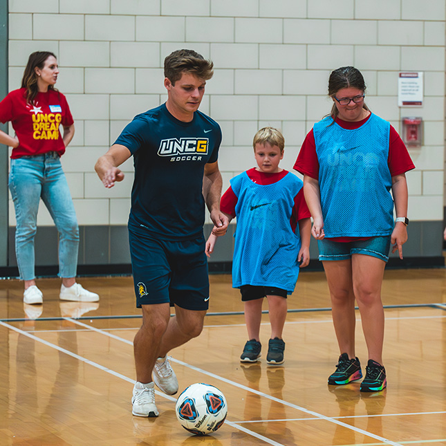 UNCG soccer player kicks a ball in a gym with 2 kids in dream camp t-shirts.