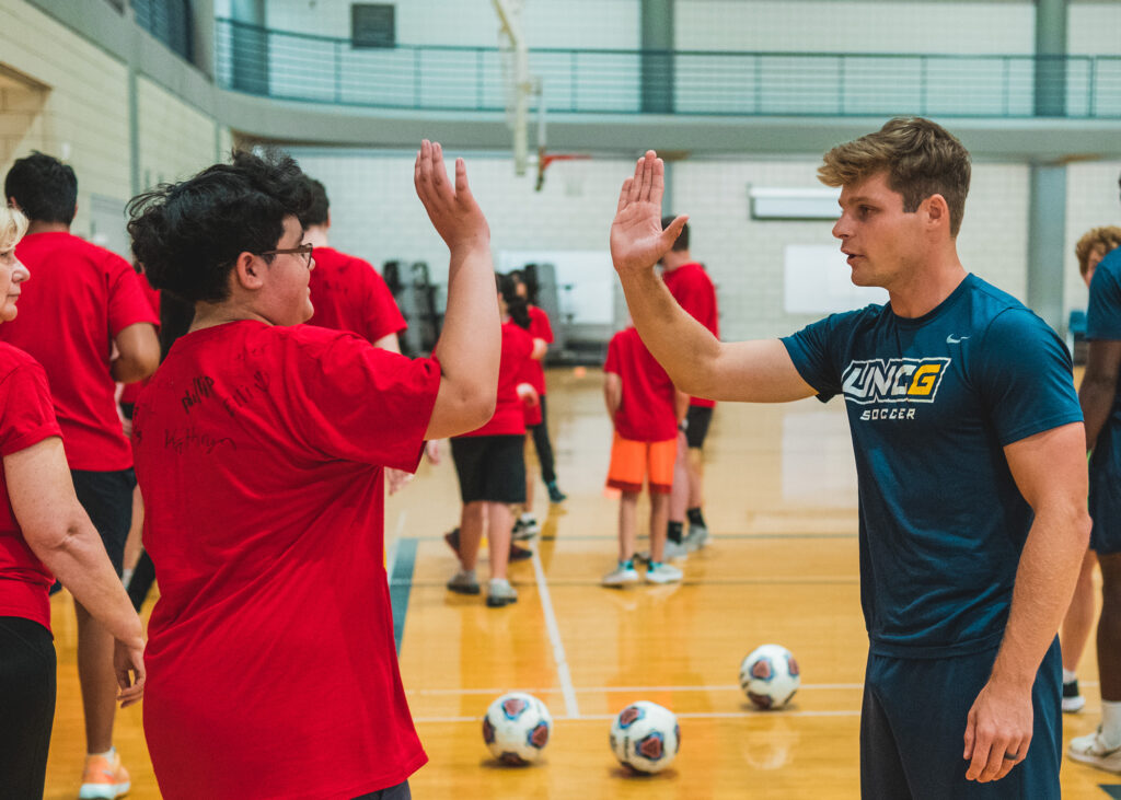 Young man high fives a teenager in a gym with soccer balls in the background. 