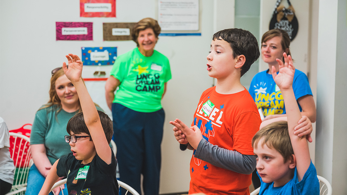 Youth in Dream Camp t-shirts raise hands to participate in a classroom while counselors look on smiling.