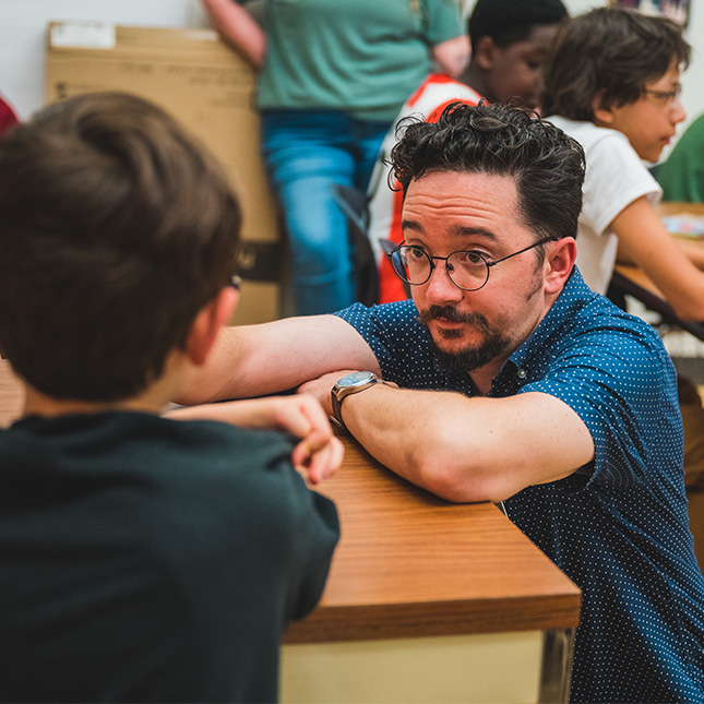 Professor kneels to talk to a young boy at a desk in a classroom.