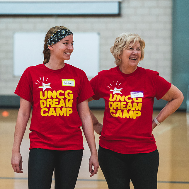 Two women in Dream Camp t-shirts smile in a gym. 