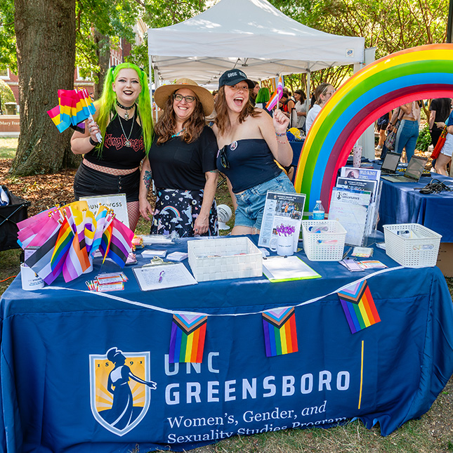 Table with rainbow flags and a UNCG women & gender studies tablecloth with 3 women posing behind it.