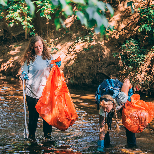Two women walk in a creek with trash bags and trash grabbing sticks.