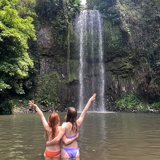 2 girls standing in water with hands raised to a waterfall. 