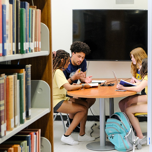 Students study at a table with bookshelves in the foreground.
