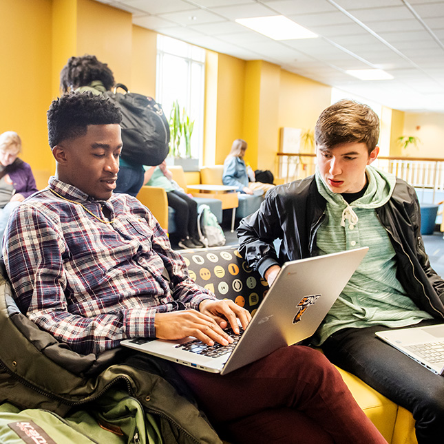 Two students study together with laptops in a lounge.