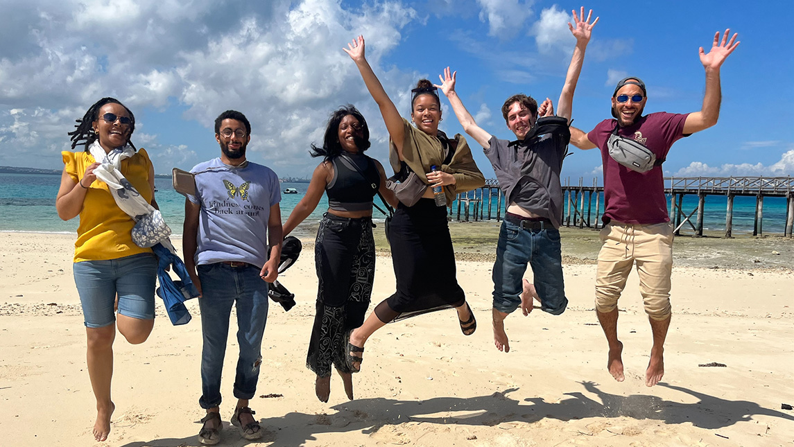 Six college students jump excitedly on a beach with a pier in the background.