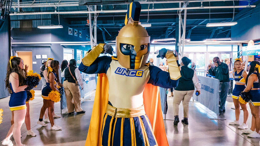 The UNCG Spiro mascot flexes his muscles around cheerleaders at a basketball stadium.