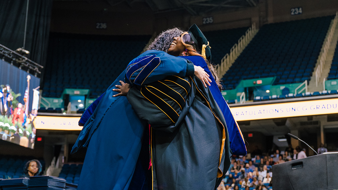 A UNCG graduate hugs her mentor on stage after receiving her doctoral hood.