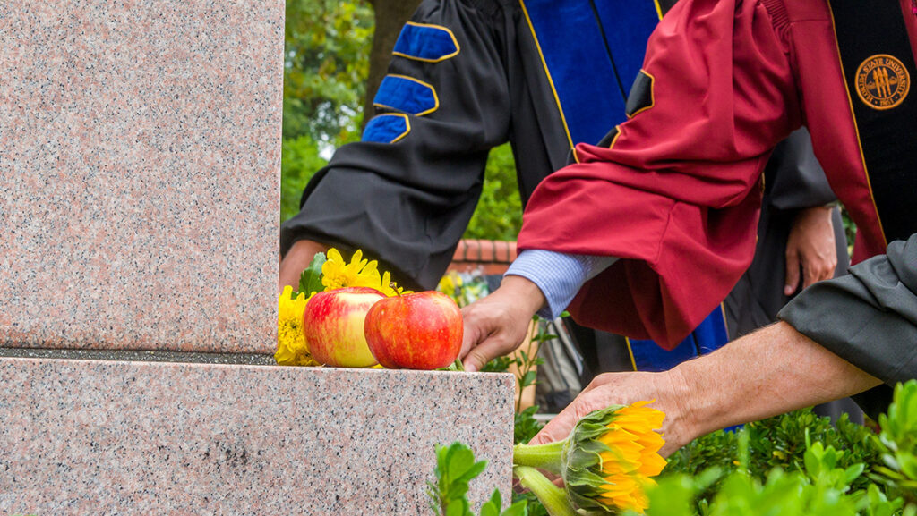 Close-up on the hands of UNCG faculty in their academic robes touching the Minerva statue base.