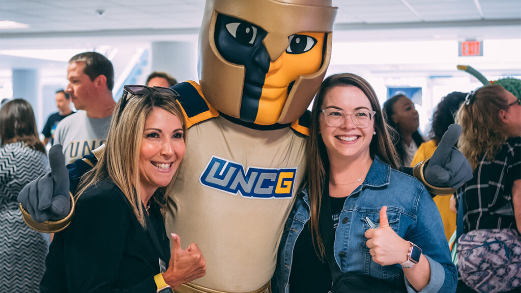 Two UNCG employees give a thumbs up while taking a picture with the mascot Spiro.
