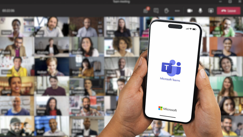 People's faces appear in boxes in a video chat on a computer screen while a person holds up their phone.