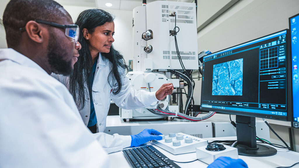 A UNCG faculty member and student in lab coats study data on a computer.