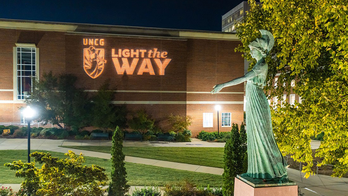 The words "Light the Way" are projected against the side of a building near the UNCG Minerva statue.