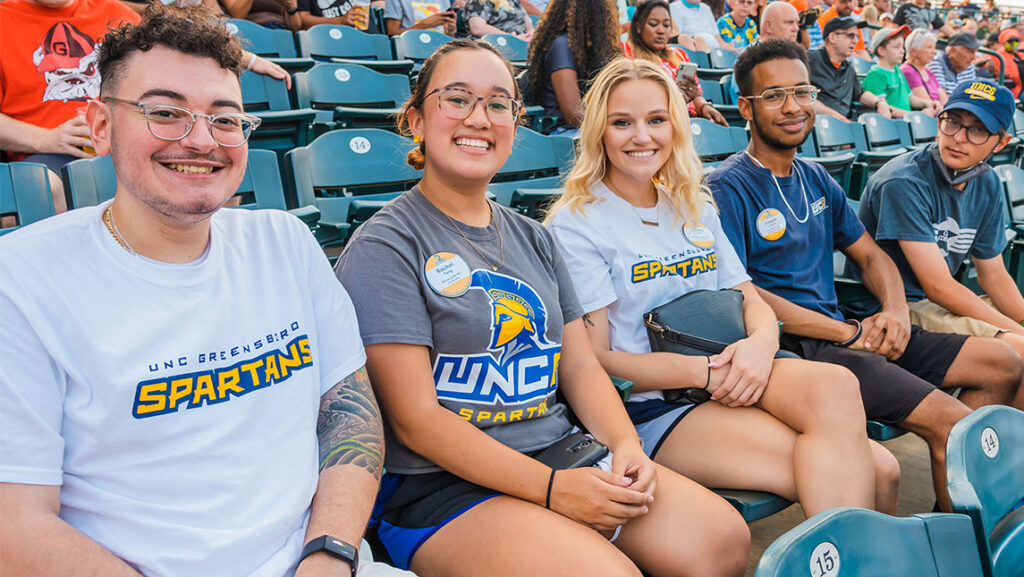 Five UNCG students sit together in the stands of a baseball stadium.