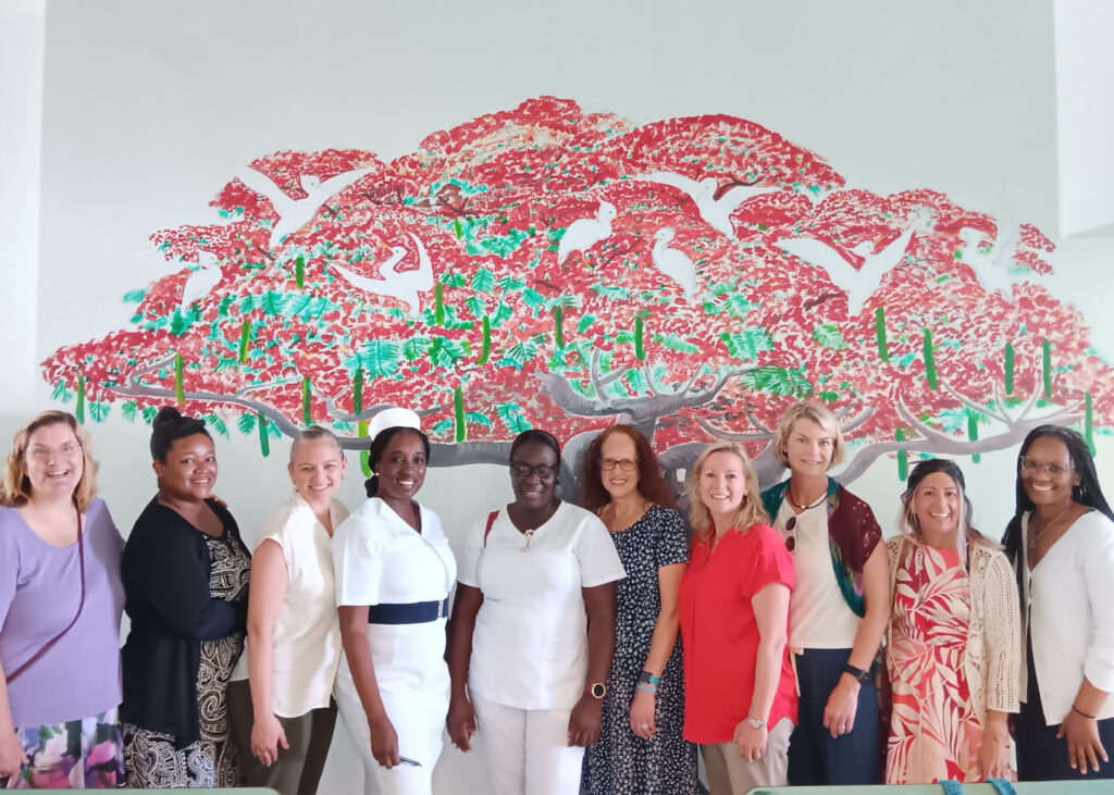 Women - some in nursing uniforms stand in front of a mural on a white wall.
