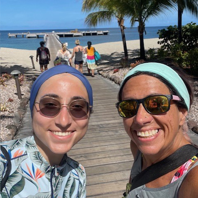 Selfie of 2 women with a pier behind them and 4 others walking out over sandy beaches and blue water with palm trees.