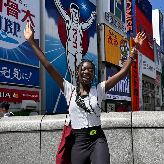 Woman poses in front of ads with Japanese characters and imitates the illustration of a runner with her hands in the air.